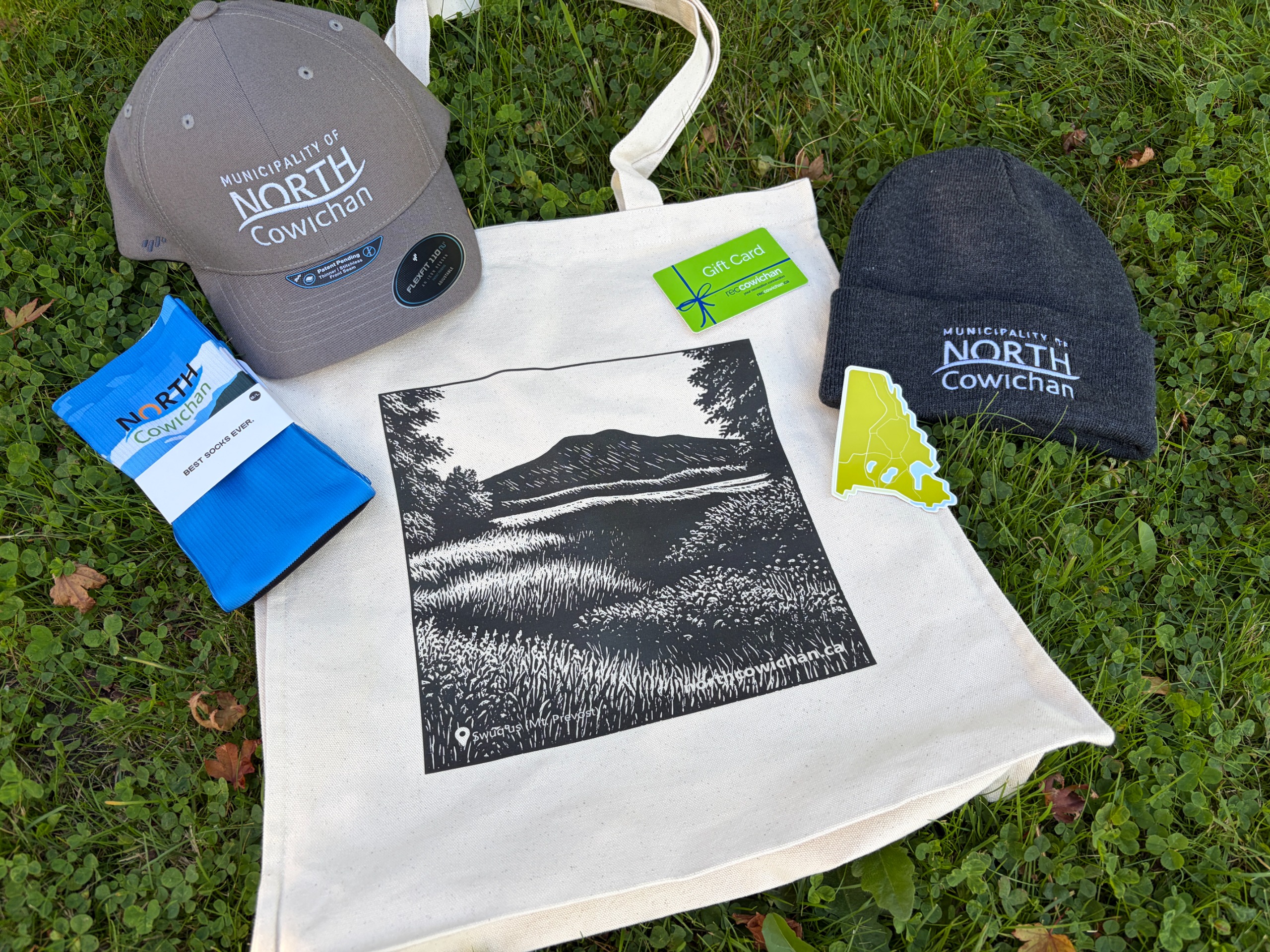 North Cowichan prizes laying on grass including a tote bag, socks, cap, toque, and recreation gift card